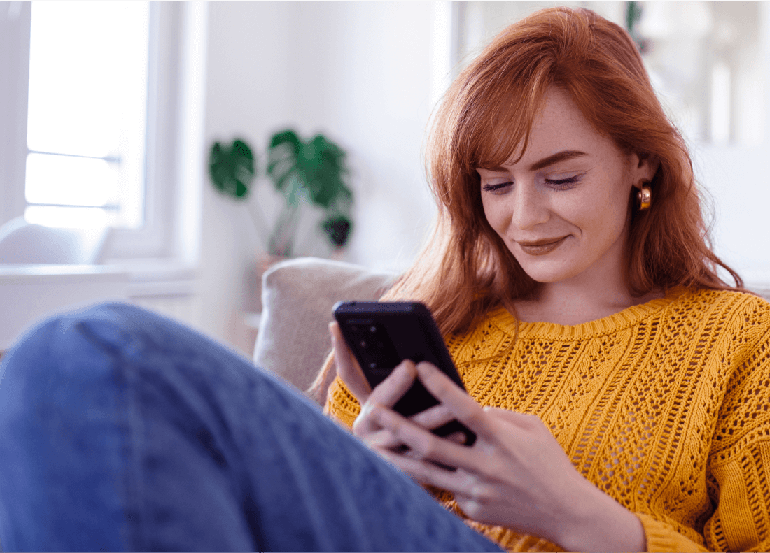 woman with red hair sitting on couch and looking at her smartphone