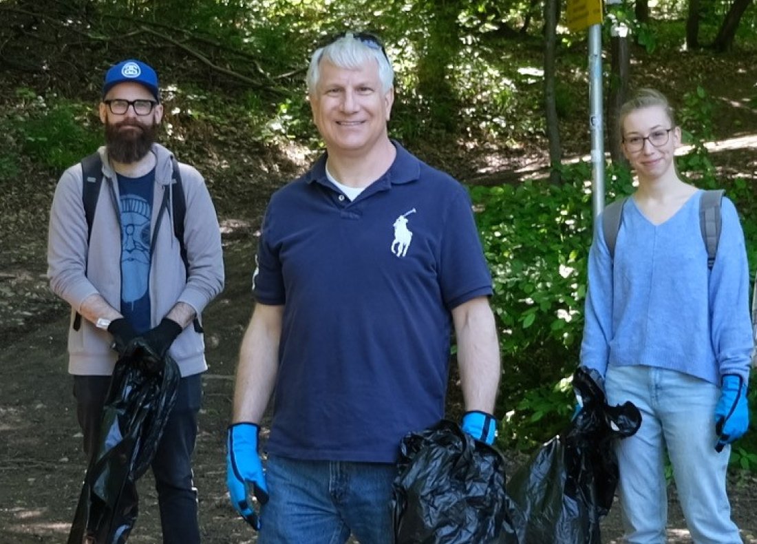 a group of people (two men and a woman) pose smiling for a picture in the forest while carrying a garbage bag and wearing protective gloves